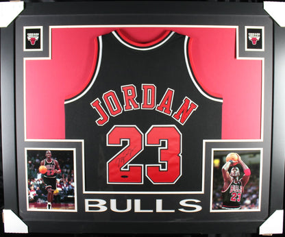 Michael Jordan signed black Chicago Bulls number 23 Mitchell and Ness jersey in a black custom frame with red matting, signed in black marker on the red numerals with the Upper Deck hologram, two action photographs below