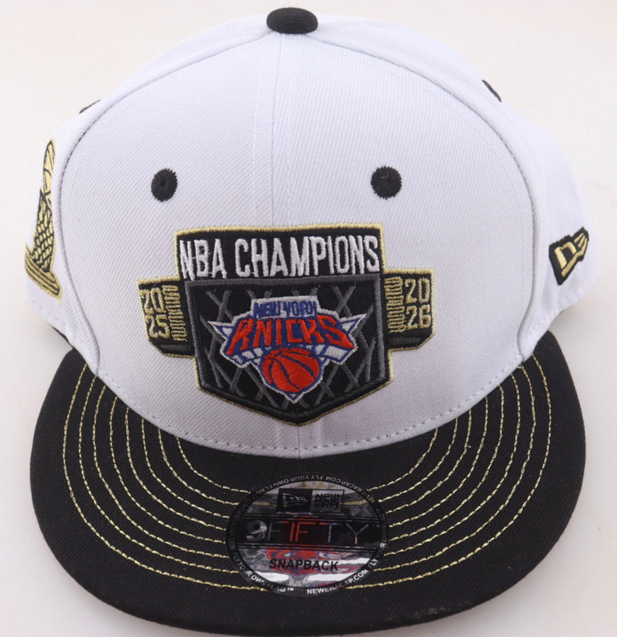 Front of the white and black New York Knicks 2025-26 NBA Champions New Era 9FIFTY snapback with the embroidered championship crest and gold stitched flat visor